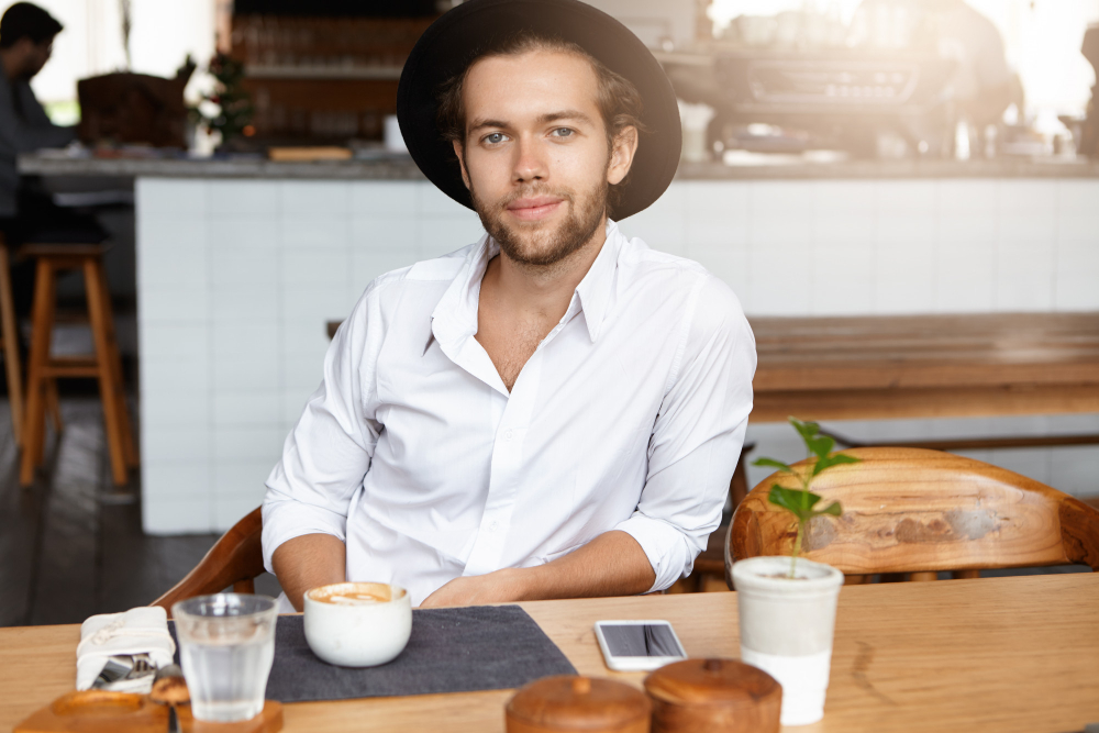 happy-cheerful-young-bearded-man-stylish-headwear-having-coffee-sitting-wooden-table-modern-cafe-interior-waiting-his-girlfriend-planning-propose-her-this-sunny-day